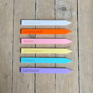 Op de afbeelding: Een verzameling kleurrijke tuinplantenmarkers in wit, oranje, roze, geel, blauw en paars. Elke marker heeft een andere kruidnaam erop gedrukt: Basilicum, Lavas, Salie, Citroengras, Oregano en Lavendel. De markers zijn gerangschikt op een houten oppervlak.