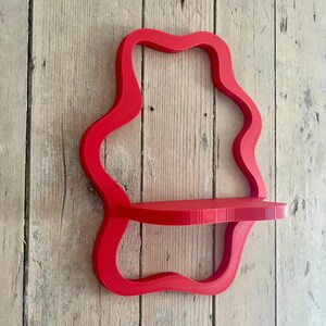 May include: A vibrant red, wavy-edged wall shelf. The shelf has a single, rectangular platform supported by a continuous, flowing frame. The shelf is mounted on a light-colored, wooden plank wall.
