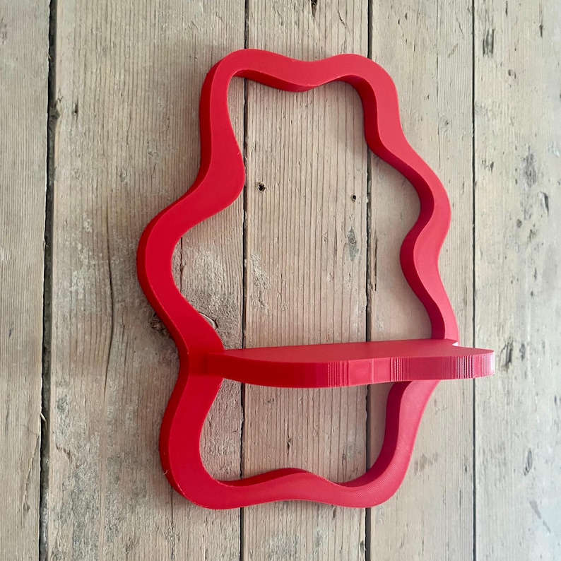 May include: A vibrant red, wavy-edged wall shelf. The shelf has a single, rectangular platform supported by a continuous, flowing frame. The shelf is mounted on a light-colored, wooden plank wall.