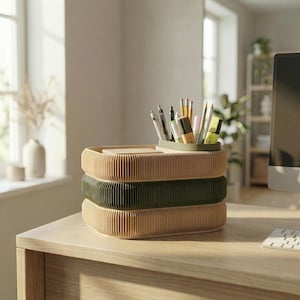 May include: A three-tiered desk organizer with a corrugated design. The top and bottom tiers are beige, and the middle tier is olive green. A small, olive green tray on top holds pens, pencils, and markers. The organizer sits on a wooden desk.