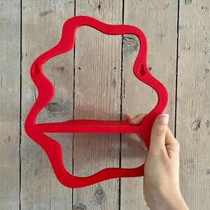May include: A bright red, wall-mounted shelf with a unique, flowing shape. The shelf has a single horizontal surface for displaying items and is held by a hand against a wooden background. The shelf is approximately 12 inches wide.