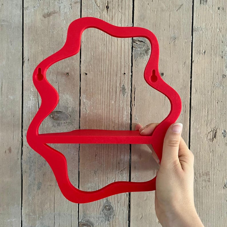 May include: A bright red, wall-mounted shelf with a unique, flowing shape. The shelf has a single horizontal surface for displaying items and is held by a hand against a wooden background. The shelf is approximately 12 inches wide.