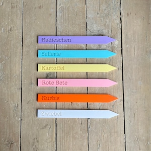 Puede incluir: Seis marcadores de jardín coloridos en lavanda, turquesa, amarillo, rosa, naranja y blanco. Cada marcador tiene una punta y texto en alemán: Radieschen, Sellerie, Kartoffel, Rote Bete, Kürbis y Zwiebel. Dispuestos sobre una superficie de madera.