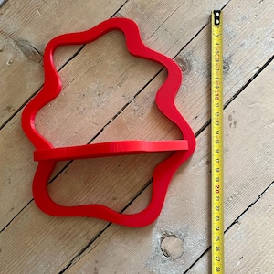 May include: A bright red, wall-mounted shelf with a unique, flowing shape. The shelf has a single horizontal surface for displaying items. A measuring tape shows the shelf is approximately 10 inches wide.