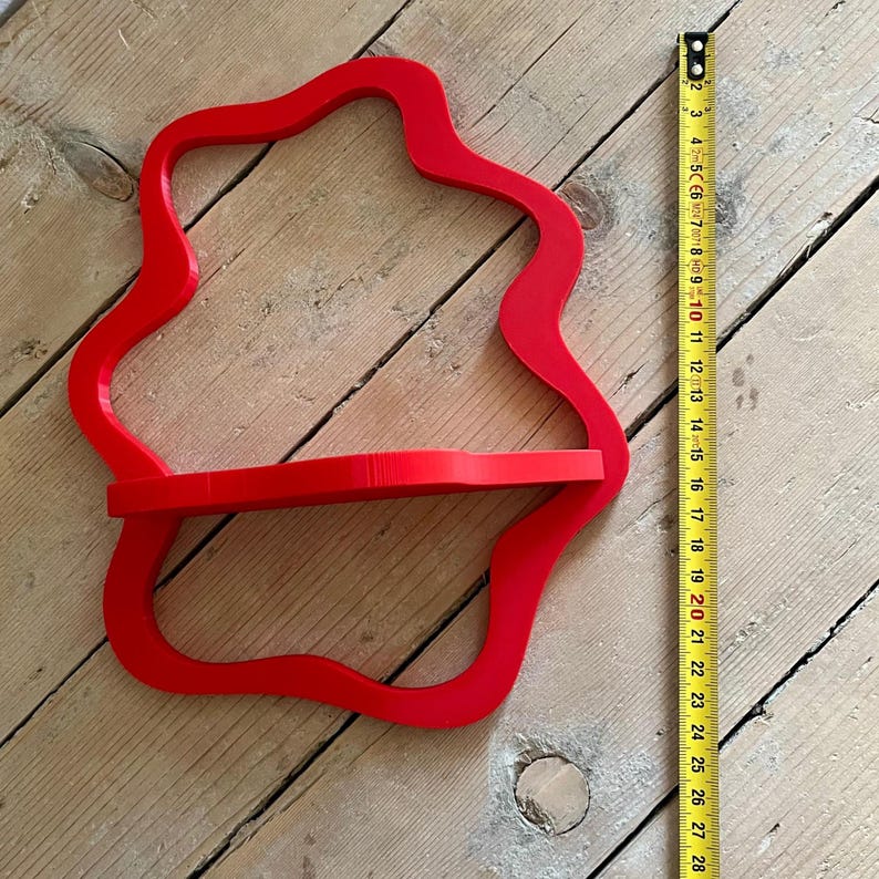 May include: A bright red, wall-mounted shelf with a unique, flowing shape. The shelf has a single horizontal surface for displaying items. A measuring tape shows the shelf is approximately 10 inches wide.