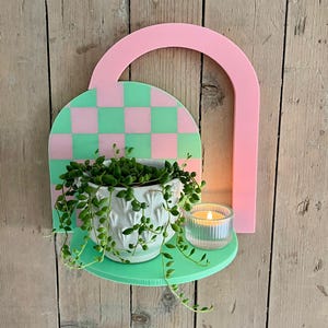 Puede incluir: Estantería decorativa de pared en colores pastel. Presenta un arco rosa, un fondo a cuadros verdes y rosas, y una balda verde. Una maceta de cerámica blanca con una planta colgante y una vela pequeña en un vaso están en la balda.