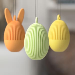 Könnte beinhalten: Drei hängende Osterschmuckstücke in Orange, Grün und Gelb. Das orangefarbene Schmuckstück hat die Form eines Hasen mit vertikalen Rillen. Die grünen und gelben Schmuckstücke sind eiförmig mit vertikalen Rillen. Alle haben eine Schlaufe zum Aufhängen.