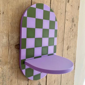 May include: A purple and green checkered wall shelf with a rounded top and a small shelf. The shelf is mounted on a wooden surface. The shelf is approximately 10 inches wide and 6 inches deep.