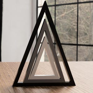 Könnte beinhalten: Eine moderne geometrische Skulptur mit drei ineinander verschachtelten Dreiecken. Das äußere Dreieck ist schwarz, das mittlere grau und das innerste weiß. Die Skulptur steht auf einer Holzoberfläche, mit einem Fenster und Grün in der Hintergrund.