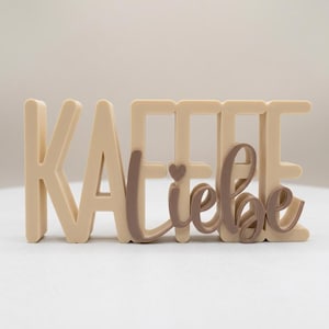 Könnte beinhalten: Ein beiges Dekoschild mit dem Wort "KAFFEE" in Blockbuchstaben und dem Wort "Liebe" in einer dunkelbraunen Schreibschrift. Ein kleines Herz ist über dem Buchstaben "i" in "Liebe" zu sehen.