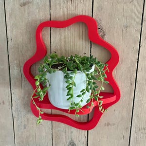May include: A red, star-shaped wall shelf holding a white ceramic pot with a trailing green plant. The shelf is mounted on a wooden plank wall, creating a decorative display for indoor plants.