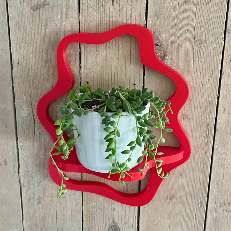 May include: A red, star-shaped wall shelf holding a white ceramic pot with a trailing green plant. The shelf is mounted on a wooden plank wall, creating a decorative display for indoor plants.