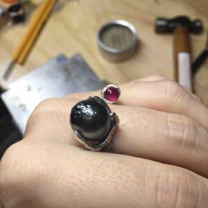 Può includere: Un anello fatto a mano con una grande gemma nera lucida come punto focale. Una gemma rossa rotonda più piccola è posta sopra la pietra nera. L'anello è incastonato in metallo argentato con un design intricato e strutturato.