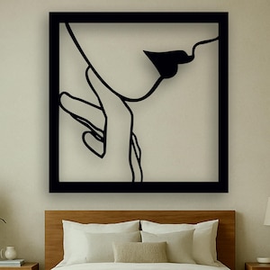 Op de afbeelding: Een zwart omlijst kunstwerk aan de muur met een minimalistische lijntekening van een gezicht, lippen en een hand. Het kunstwerk hangt boven een houten bed met wit beddengoed, een beige vloerkleed en twee houten nachtkastjes.