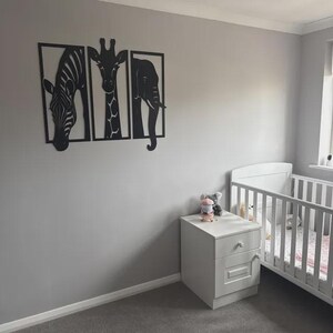 May include: Black metal wall art set featuring a zebra, giraffe, and elephant, each framed in a rectangular shape. The artwork is mounted on a gray wall in a nursery setting, with a white crib and nightstand.