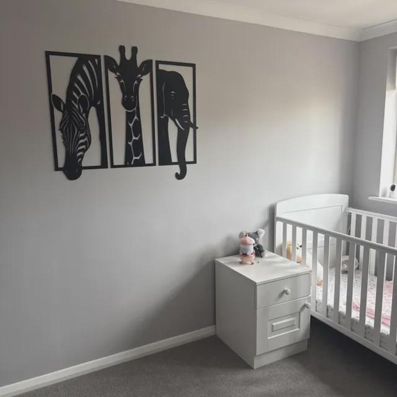 May include: Black metal wall art set featuring a zebra, giraffe, and elephant, each framed in a rectangular shape. The artwork is mounted on a gray wall in a nursery setting, with a white crib and nightstand.