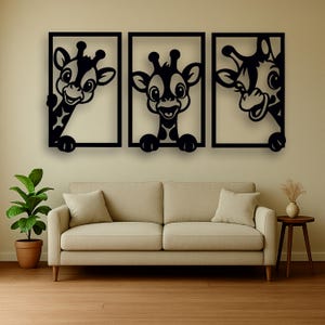 May include: Three black framed wall art pieces featuring cartoon giraffes. Each giraffe has a smiling face and is peeking out from behind the frame. The artwork is displayed above a beige sofa in a living room setting.