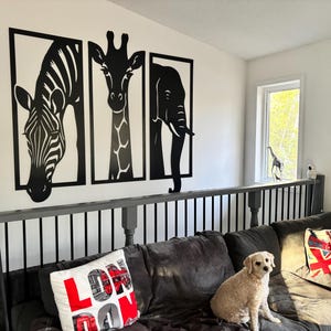 May include: Black and white wall art featuring a zebra, giraffe, and elephant. The animal silhouettes are framed and mounted on a white wall. A dog sits on a dark gray sofa with decorative pillows. A framed picture and a window are also visible.