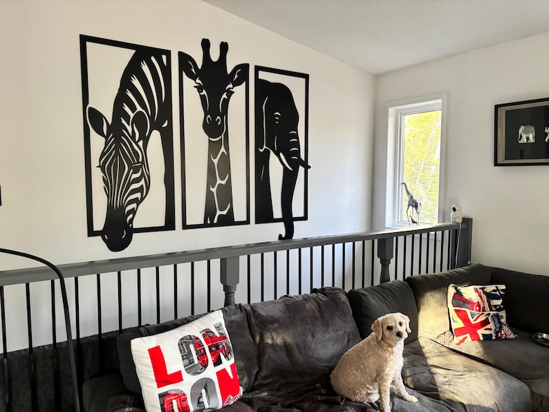 May include: Black and white wall art featuring a zebra, giraffe, and elephant. The animal silhouettes are framed and mounted on a white wall. A dog sits on a dark gray sofa with decorative pillows. A framed picture and a window are also visible.