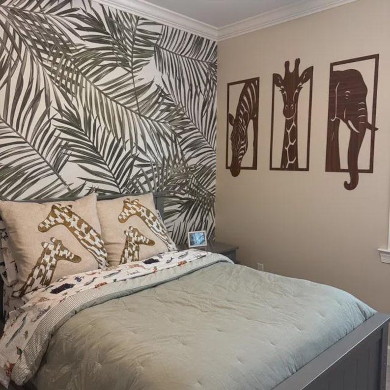 May include: A bedroom with a gray bed frame, sage green bedding, and giraffe-patterned pillows. The wall features a tropical leaf wallpaper and wooden animal cutouts of a zebra, giraffe, and elephant.