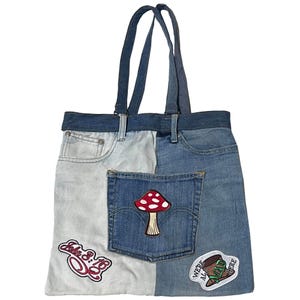 May include: A patchwork denim tote bag with a light blue and medium blue color scheme. The bag features a mushroom patch, a "John 3:16" patch, and a "We're All Mad Here" patch. The bag has two denim handles.
