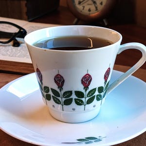 Pu&ograve; includere: Una tazza e un piattino bianchi con un motivo floreale a rose. La tazza &egrave; riempita con un liquido scuro, appoggiata su un piattino bianco. Un orologio vintage e libri sullo sfondo suggeriscono un'atmosfera accogliente.