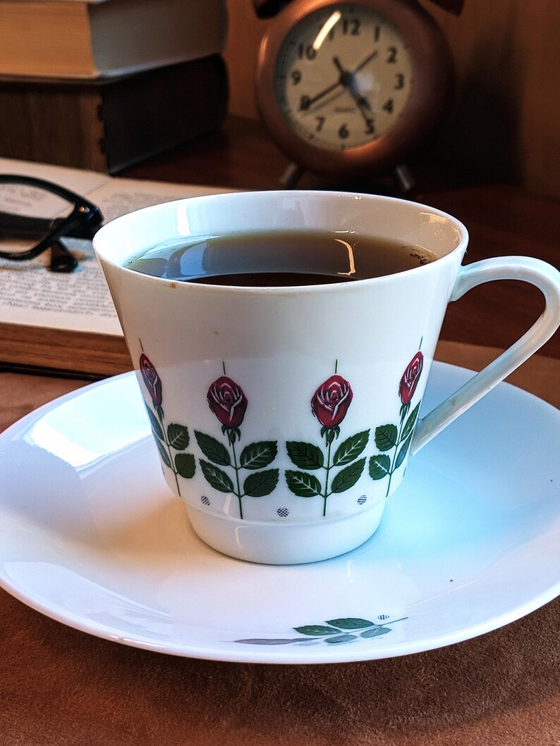 Pu&ograve; includere: Una tazza e un piattino bianchi con un motivo floreale a rose. La tazza &egrave; riempita con un liquido scuro, appoggiata su un piattino bianco. Un orologio vintage e libri sullo sfondo suggeriscono un'atmosfera accogliente.