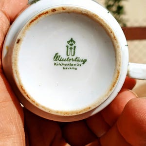 Pu&ograve; includere: Base di una tazza da t&egrave; in porcellana bianca con logo verde e scritta "Wieslerlissg Kirchenlamitz BAYERN". La tazza ha un bordo marrone e un piccolo manico.