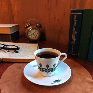 Pu&ograve; includere: Una tazza di caff&egrave; su un piattino con un motivo floreale, appoggiata su un tappetino di pelle marrone. Una sveglia vintage in rame, libri e occhiali sono sullo sfondo. La tazza ha un motivo a rose.