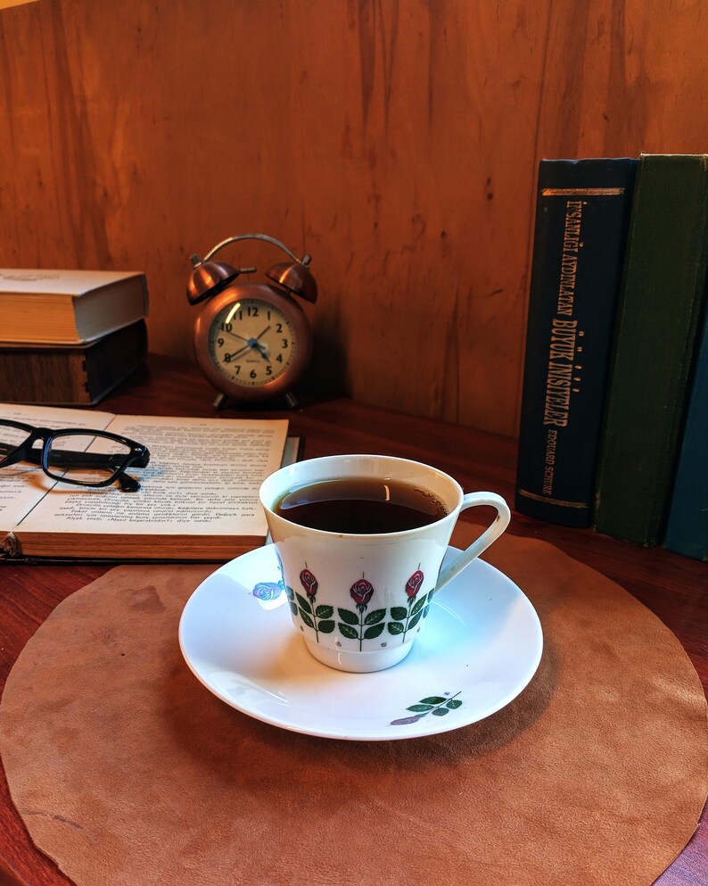 Pu&ograve; includere: Una tazza di caff&egrave; su un piattino con un motivo floreale, appoggiata su un tappetino di pelle marrone. Una sveglia vintage in rame, libri e occhiali sono sullo sfondo. La tazza ha un motivo a rose.