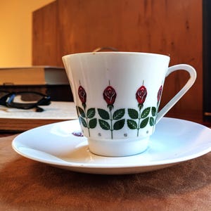 Pu&ograve; includere: Un set di tazza e piattino bianchi con un motivo a rose. La tazza presenta rose rosse con foglie e steli verdi. Il piattino &egrave; bianco e rotondo. Il set &egrave; su una superficie marrone.