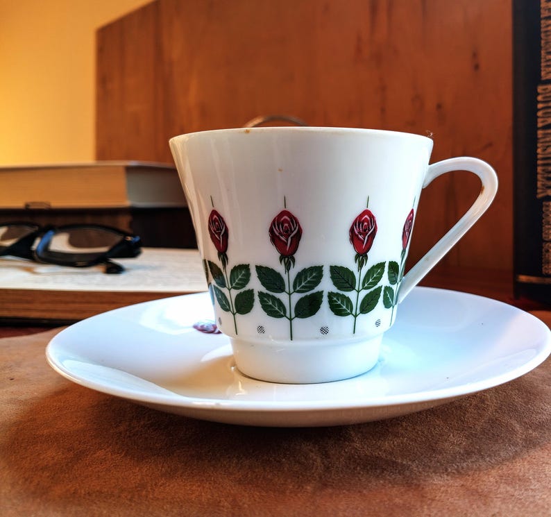 Pu&ograve; includere: Un set di tazza e piattino bianchi con un motivo a rose. La tazza presenta rose rosse con foglie e steli verdi. Il piattino &egrave; bianco e rotondo. Il set &egrave; su una superficie marrone.