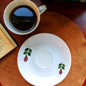 Pu&ograve; includere: Una tazza bianca piena di liquido scuro &egrave; accanto a un piattino abbinato. Il piattino &egrave; decorato con una rosa rossa e foglie verdi. La tazza e il piattino sono su una superficie marrone.