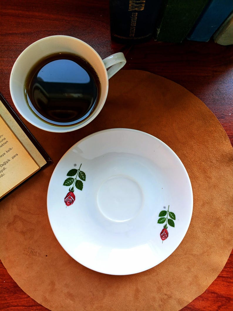 Pu&ograve; includere: Una tazza bianca piena di liquido scuro &egrave; accanto a un piattino abbinato. Il piattino &egrave; decorato con una rosa rossa e foglie verdi. La tazza e il piattino sono su una superficie marrone.