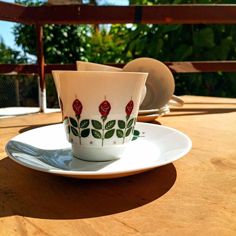 Pu&ograve; includere: Servizio da t&egrave; bianco con motivo a rose rosse e foglie verdi. La tazza &egrave; appoggiata su un piattino bianco. Altre tazze e vegetazione sono visibili su una superficie di legno sullo sfondo.