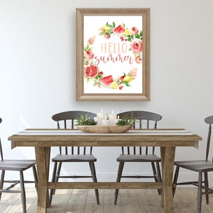 May include: A framed watercolor print with a wreath of summer fruits and flowers. The wreath surrounds the text "HELLO SUMMER". The print is hanging on a white wall above a wooden table with four chairs.