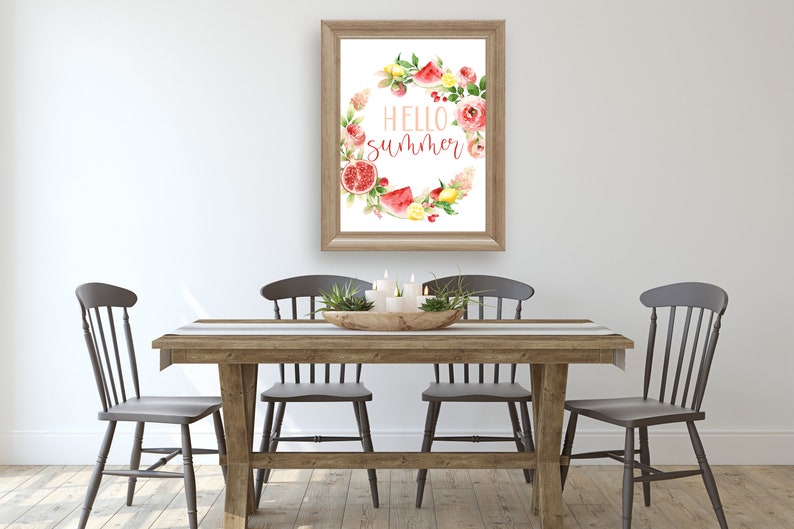 May include: A framed watercolor print with a wreath of summer fruits and flowers. The wreath surrounds the text "HELLO SUMMER". The print is hanging on a white wall above a wooden table with four chairs.