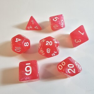 May include: A collection of seven red polyhedral dice with white numbering. The dice include a tetrahedron, a cube, a d8, a d10, and a d20, suitable for tabletop role-playing games.