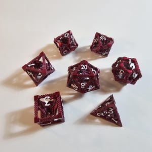 Pode incluir: Conjunto de sete dados poliédricos bordô para jogos de tabuleiro. Cada dado apresenta um design detalhado e vazado com numeração branca. O conjunto inclui um D4, D6, D8, D10, D12, D20 e um dado percentual.