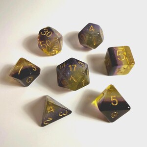 May include: A set of seven polyhedral dice featuring a gradient of translucent yellow and deep purple resin with embedded glitter. Each die has engraved numbers filled with gold ink, including a d20, d12, d10, d8, d6, d4, and a percentile die.