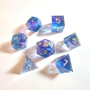 Op de afbeelding: Set van zeven polyhedrale dobbelstenen in blauwe en paarse tinten. Elke dobbelsteen is transparant met ingebedde decoratieve elementen en zilveren cijfers. De dobbelstenen zijn gerangschikt op een wit oppervlak.
