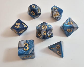 May include: A set of seven polyhedral dice in shades of blue, white, and gold. The dice are arranged on a white surface. Each die has gold numbering. The set includes a D4, D6, D8, D10, D12, D20, and a percentile die.