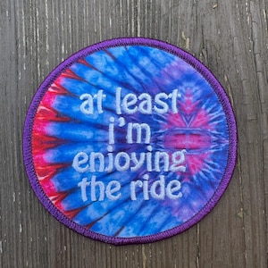 May include: A circular embroidered patch featuring a vibrant blue, purple, and red tie-dye pattern. The text "at least i'm enjoying the ride" is stitched in white thread across the center. The patch has a dark purple stitched border.