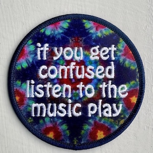 May include: Round patch with a tie-dye design in shades of blue, red, and yellow. The patch has a navy blue border and white embroidered text that reads "if you get confused listen to the music play."