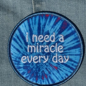 May include: A circular embroidered patch featuring a blue and dark blue tie-dye pattern with red accents. The text "i need a miracle every day" is stitched in white thread across the center of the patch, which has a dark blue stitched border.