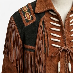 K&ouml;nnte beinhalten: Eine braune Wildlederjacke mit langen Fransen, schwarzen Wildlederschulterpartien mit Perlenakzenten und wei&szlig;en, knochenartigen R&ouml;hrenperlen in horizontalen Reihen entlang des Vorderverschlusses, abgerundet durch eine geflochtene Lederkante.