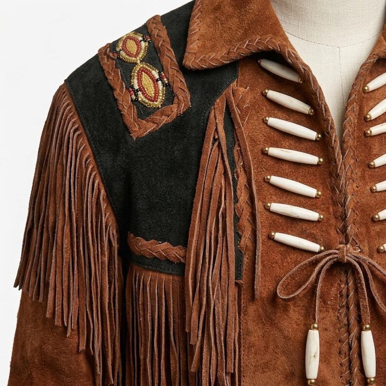 K&ouml;nnte beinhalten: Eine braune Wildlederjacke mit langen Fransen, schwarzen Wildlederschulterpartien mit Perlenakzenten und wei&szlig;en, knochenartigen R&ouml;hrenperlen in horizontalen Reihen entlang des Vorderverschlusses, abgerundet durch eine geflochtene Lederkante.
