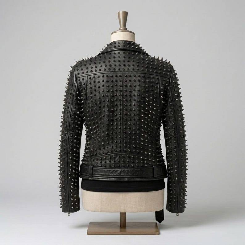 May include: A black jacket featuring a dense pattern of pointed metal studs covering the entire back and sleeves. The garment includes a structured collar, a horizontal waist belt with a buckle, and long sleeves, displayed on a mannequin torso.