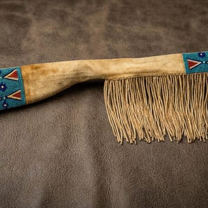 以下が含まれることがあります： ビーズのアクセントとフリンジが付いた、タン色の革製の鞘。鞘の両端には、赤と白の幾何学模様が施されたターコイズブルーのビーズパネルが付いています。フリンジは薄いタン色です。
