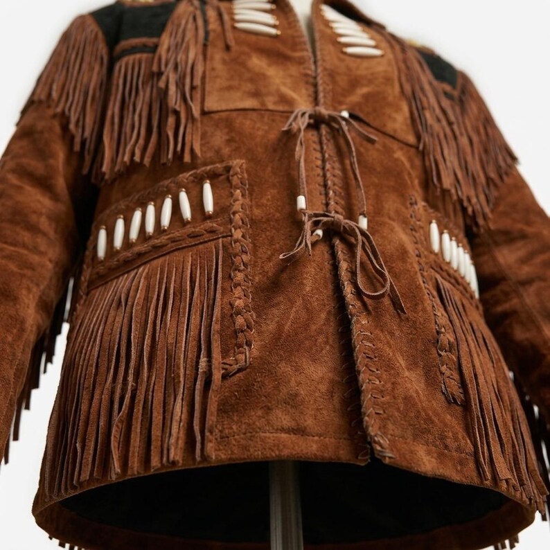 K&ouml;nnte beinhalten: Eine braune Wildlederjacke mit langen Fransen an Brust und &Auml;rmeln. Das Kleidungsst&uuml;ck ist mit wei&szlig;en zylindrischen Perlen, geflochtenen Lederverzierungen an den N&auml;hten und zwei vorderen Bindeb&auml;ndern aus d&uuml;nnen Lederriemen versehen.
